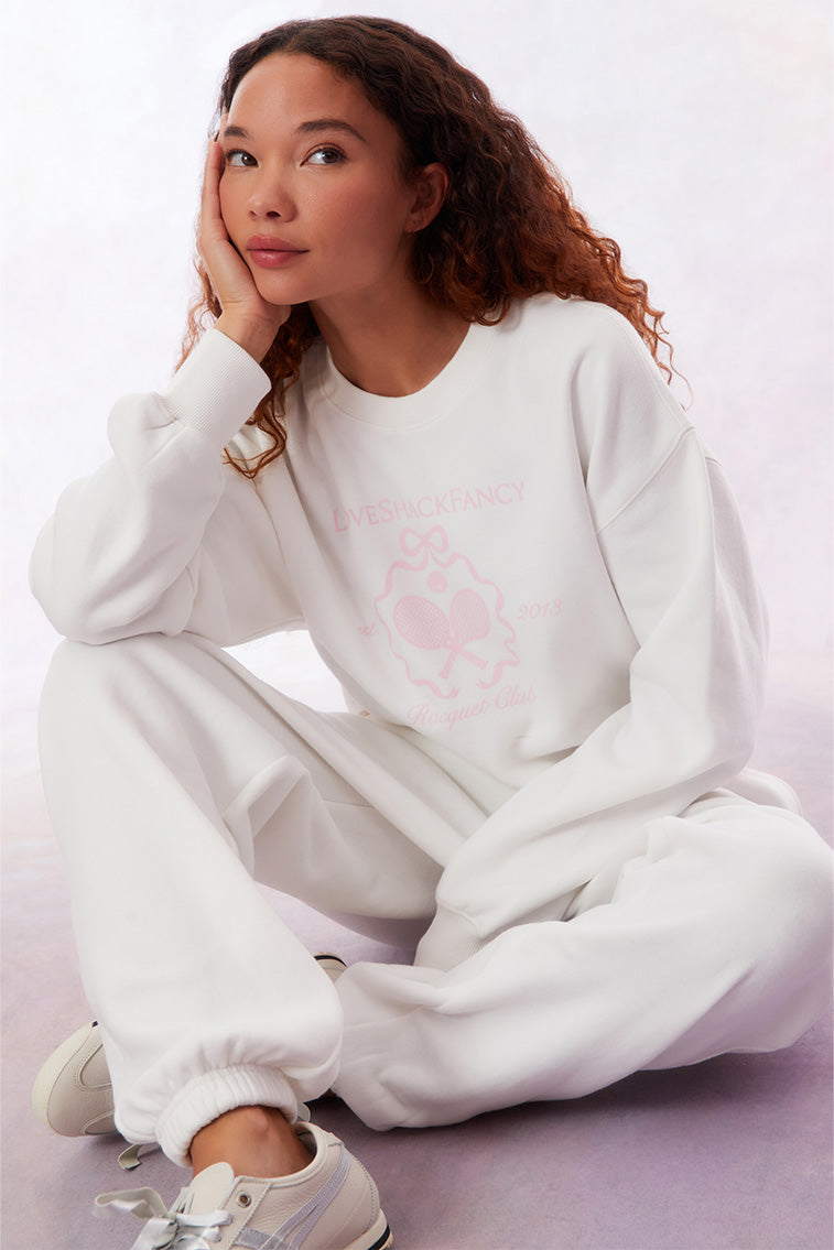 White pullover crew-neck sweater with ribbed sleeve cuffs, neckline, and hem. With a pink LoveShackFancy logo including two tennis raquets.
