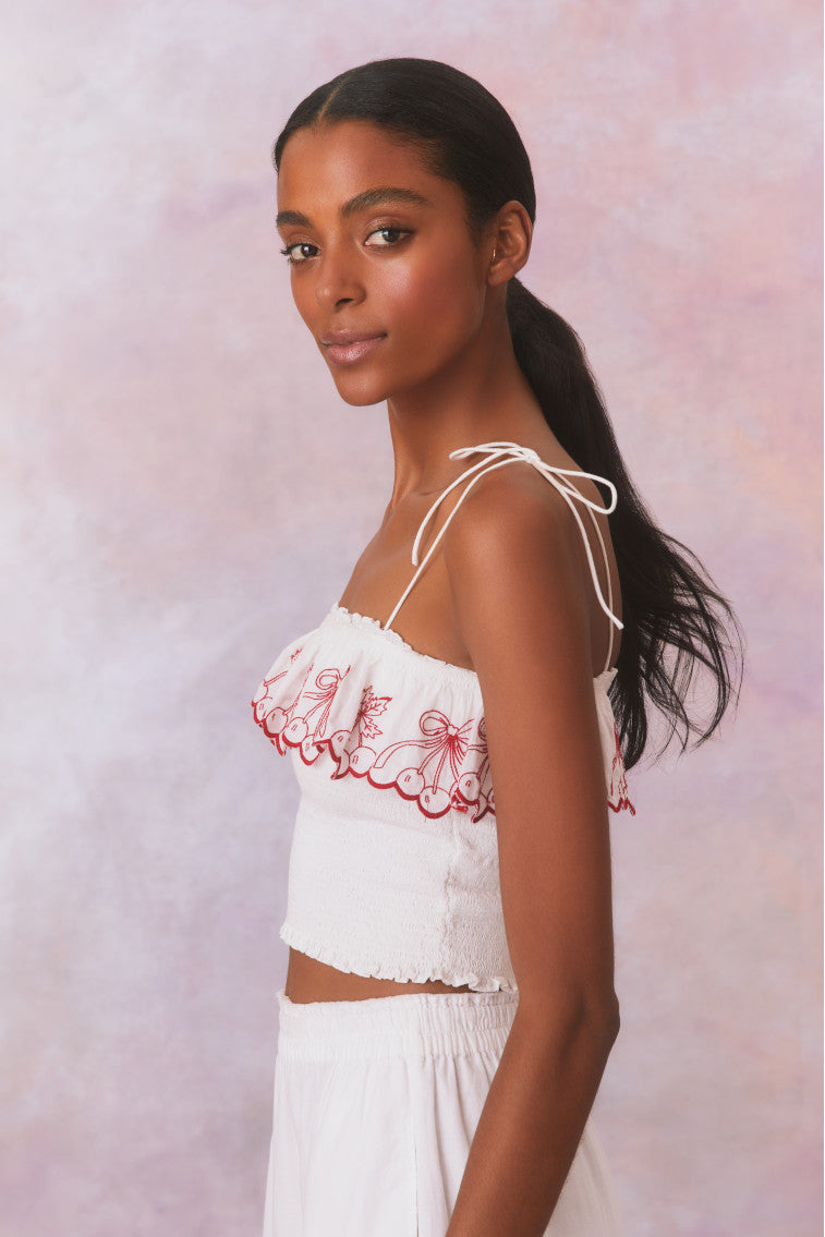 White organic cotton top with spaghetti straps that self tie. Smocked bodice with scalloped red cherry embroideries as a ruffle on the square neckline. Pairs with Amparo Skirt for a full set.