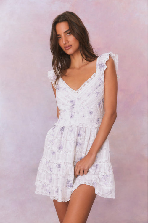 White mini dress with blue/purple floral details and an open v-neck with elasticated flutter sleeves and a smocked waist, the bodice and skirt contain lace and embroidery and is complete with pockets.