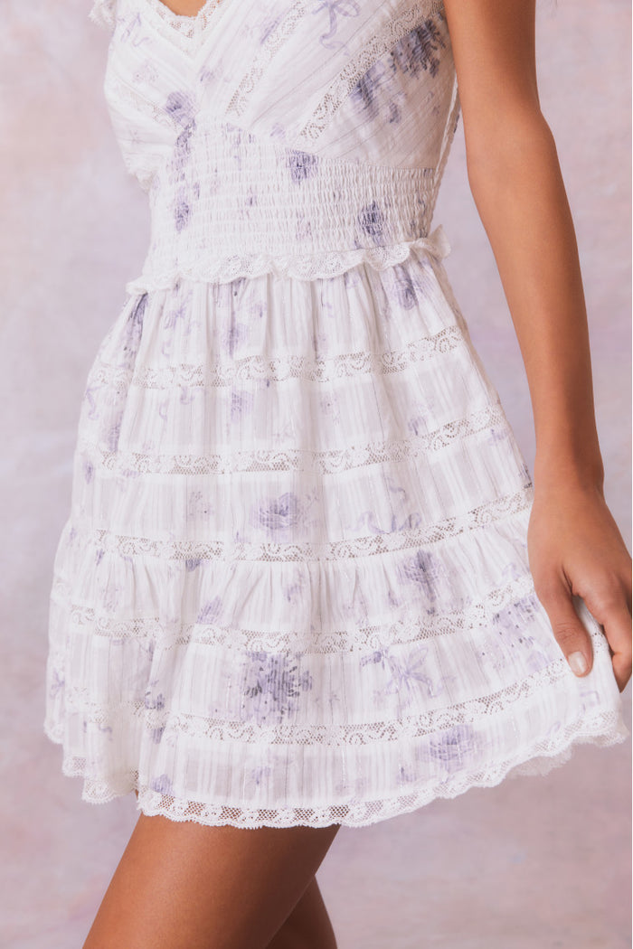 White mini dress with blue/purple floral details and an open v-neck with elasticated flutter sleeves and a smocked waist, the bodice and skirt contain lace and embroidery and is complete with pockets.